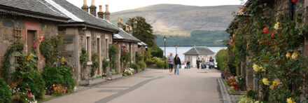 luss-village-pier-road-with-loch-lomond-and-tourists-in-the-distance-and-stone-houses-decorated-with-flowers-on-both-sides-of-the-road