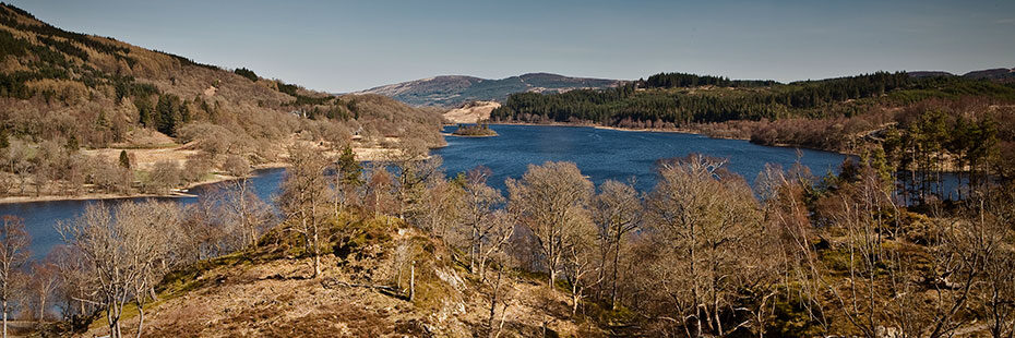 loch-achray-in-trossachs-surrounded-by-very-brown-landscape-of-bare-trees-and-coniferous-forest-in-the-distance