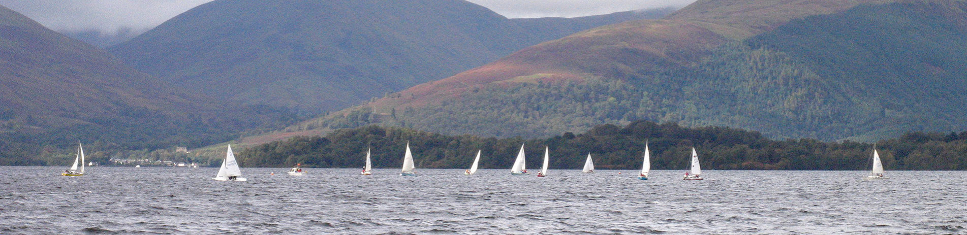 several-sailing-boats-with-white-veils-in-the-distance-on-loch-and-surrounding-high-hills-covered-by-forests-on-the-right