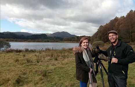 two-young-volunteers-in-jackets-at-the-edge-of-loch-venachar-one-holding-binoculars-and-with-tripod-set-up-in-front-of-them-performing-a-bird-survey