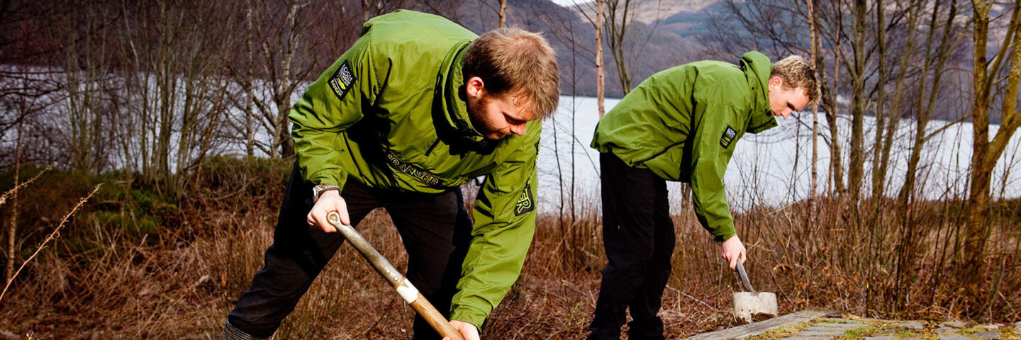 two-blond-male-rangers-in-green-jackets-digging-next-to-path-next-to-bare-trees-and-loch-in-the-distance