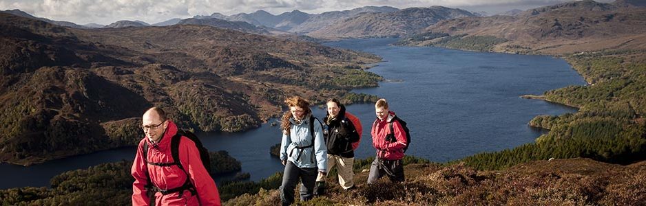 group-of-four-walkers-two-male-two-female-dressed-in-outdoor-gear-reaching-ben-aan-hill-summit-on-path-single-file-loch-katrine-and-trossachs-hills-visible-behind