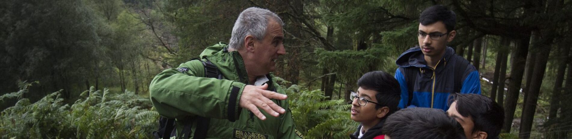 national-park-ranger-in-green-jacket-demonstrating-something-using-his-hands-in-front-of-school-students-in-the-forest