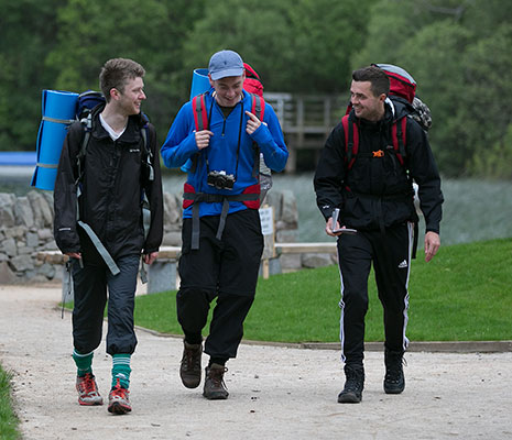 three-young-men-in-hiking-gear-and-large-backpacks-with-sleeping-mats-visible-walking-and-chatting-animatedly-on-path