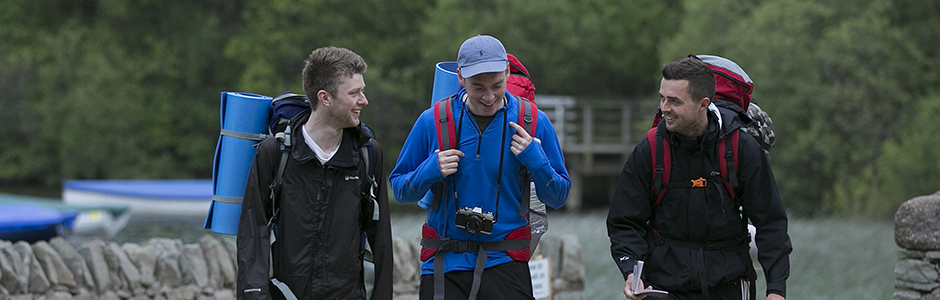 close-up-of-three-young-men-in-hiking-gear-and-large-backpacks-with-sleeping-mats-visible-walking-and-chatting-animatedly-on-path