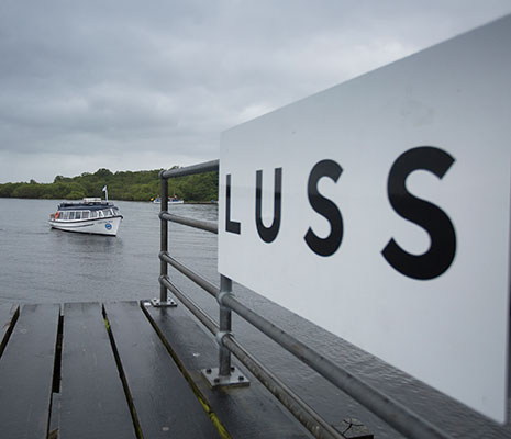 luss-pier-with-luss-name-in-close-up-letetrs-on-white-board-with-a-waterbus-on-loch-lomond-behind-approaching-the-pier