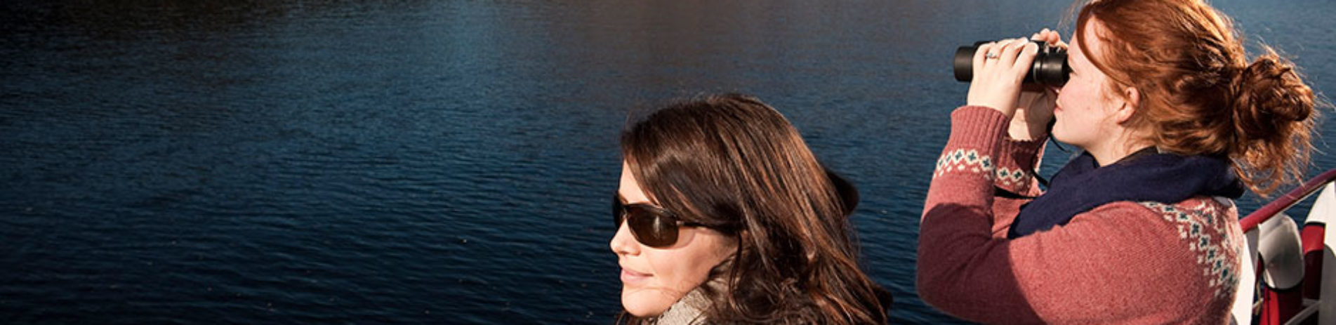 two-women-admiring-loch-lomond-from-boat-deck-one-is-looking-through-pair-of-binoculars-and-both-are-wearing-sweaters-and-woolly-scarfs