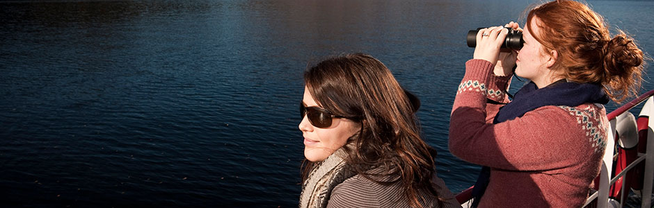 two-women-admiring-loch-lomond-from-boat-deck-one-is-looking-through-pair-of-binoculars-and-both-are-wearing-sweaters-and-woolly-scarfs