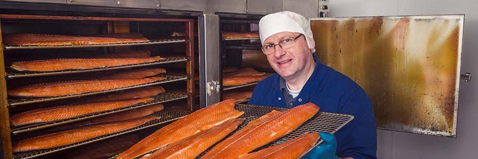 man-in-blue-t-shirt-and-protective-hat-and-glasses-holding-crate-of-smoked-salmon-next-to-container-with-more-fish-smokery-house