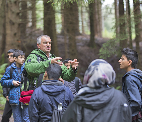 national-park-ranger-in-green-jacket-demonstrating-something-using-his-hands-in-front-of-school-students-in-the-forest