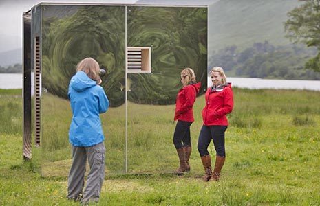 woman-in-blue-jacket-photographing-her-friend-in-red-jacket-next-to-mirrored-cabin-art-installation-in-the-middle-of-field-in-balquhidder-glen