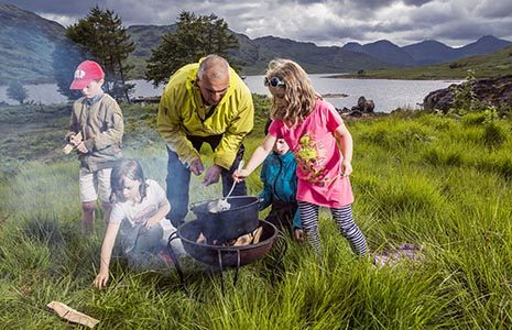 man-in-light-green-jacket-with-short-white-hair-cooking-with-four-young-children-on-grass-with-lush-loch-arklet-landscape-behind-foraging-event