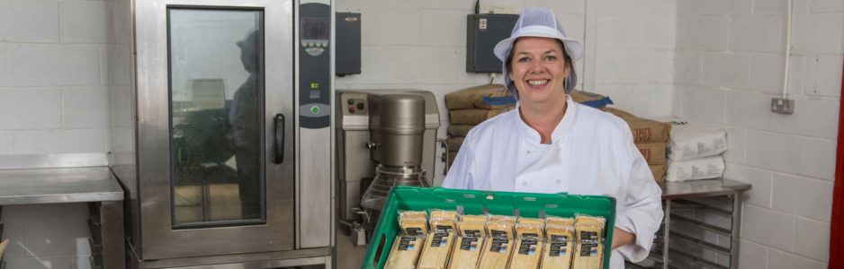 woman-in-white-protective-clothes-and-hat-in-kitchen-holding-crate-of-cheese-and-smiling-at-camera