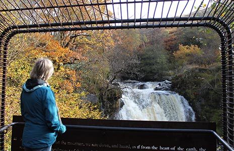 blonde-woman-in-blue-jacket-admiring-falls-of-falloch-from-woven-sound-art-installation