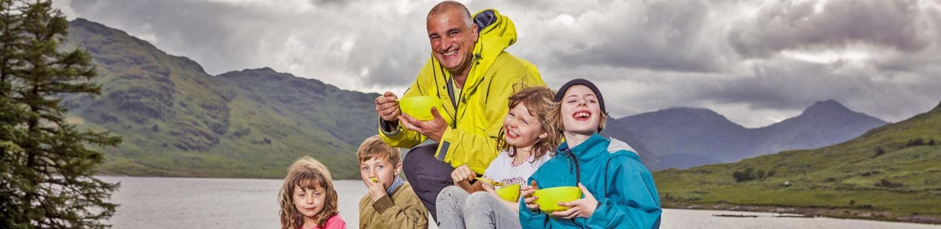 man-in-light-green-jacket-with-short-white-hair-eating-from-bowls-with-four-young-children-on-grass-with-lush-loch-arklet-landscape-behind-foraging-event