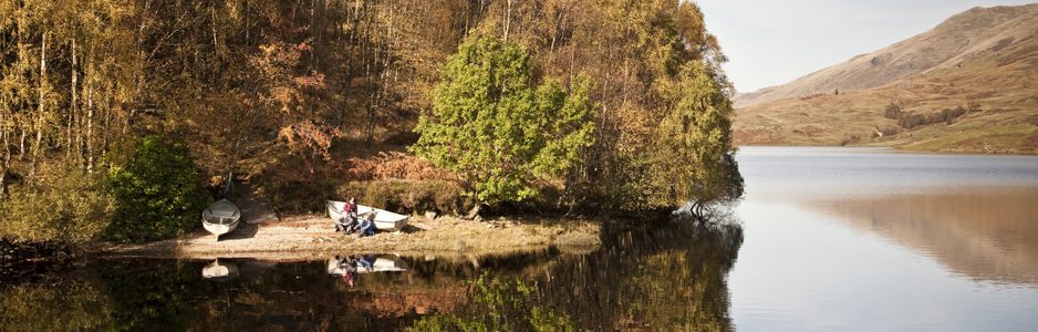 glen-finglas-reservoir-with-autumn-colours-forest-by-shore-two-boats-on-land-people-visible-next-to-one-of-them-illuminated-by-sunshine