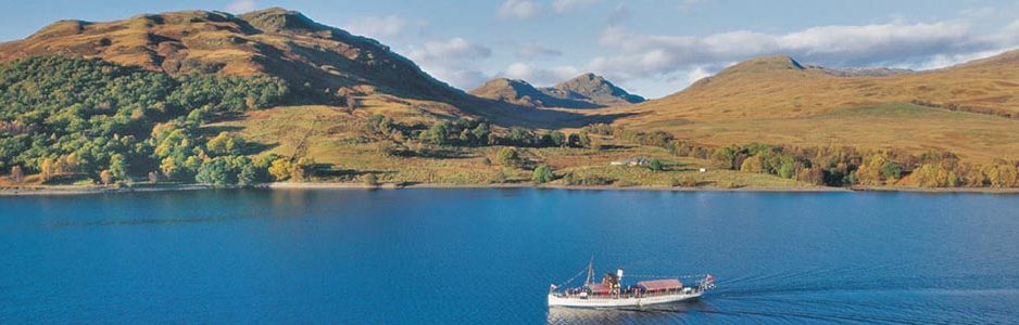 loch-katrine-blue-surface-water-crossed-by-white-steamship-hills-in-the-distance-with-ptaches-of-great-trossachs-forest-on-left