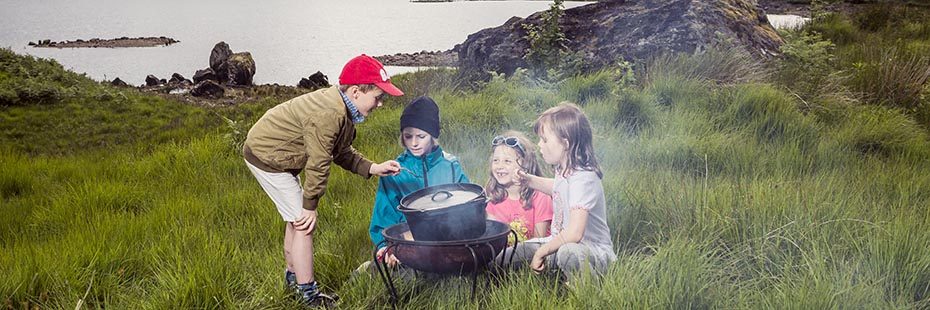 four-young-children-cooking-stooped-over-steaming-pot-on-grass-with-lush-loch-arklet-landscape-behind-foraging-event