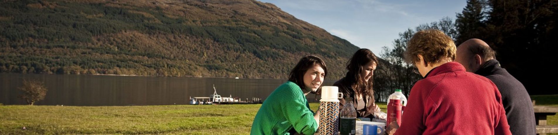 two-women-and-two-men-having-food-at-picnic-table-with-stunning-view-of-loch-and-hills-behind