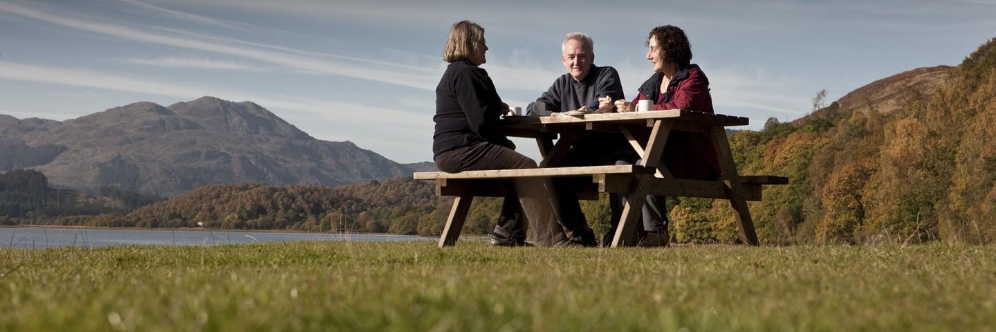 two-women-and-man-having-food-at-picnic-table-with-stunning-view-of-loch-and-hills-behind