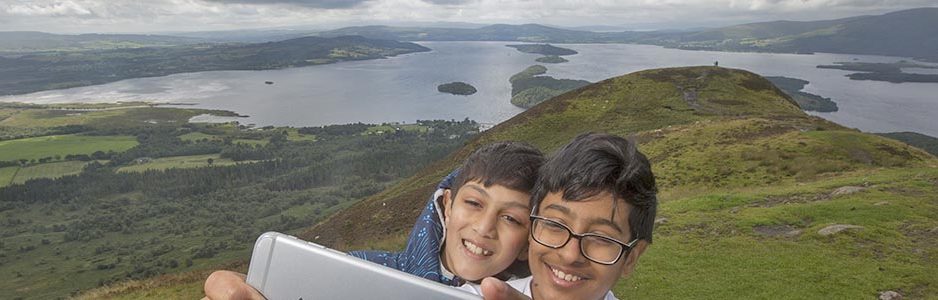 two-young-boys-smiling-and-taking-a-selfie-on-conic-hill-with-loch-lomond-behind