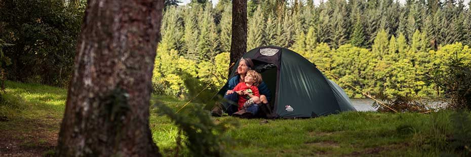 mother-holding-her-young-son-both-dressed-in-colourful-outdoor-gear-sit-at-entrance-of-dark-green-tent-looking-in-the-distance-loch-drunkie-and-coniferous-forests-in-the-sunlight-are-visible-behind-them