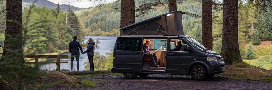 motorhome-parked-at-three-lochs-forest-drive-with-stunning-view-of-loch-and-coniferous-forests-around-the-door-is-open-and-two-men-sit-inside-one-is-reading-a-newspaper-while-a-couple-hold-hands-outside-enjoying-the-view