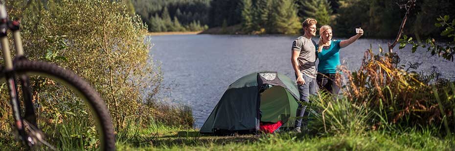 young-blond-couple-next-to-a-tent-taking-a-selfie-at-three-lochs-forest-drive-there-is-a-bike-in-the-foreground-and-loch-drunkie-and-forests-behind-them