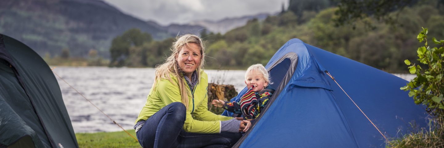 blonde-woman-holding-baby-at-blue-tent-entrance-at-edge-of-loch-chon