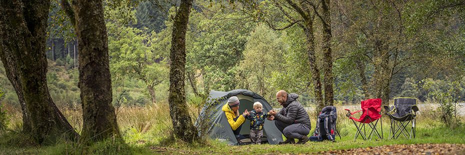 young-couple-crouched-next-to-tent-entrance-holding-baby-standing-on-his-two-feet-at-loch-chon-campsite-in-forest-folding-chairs-open-on-the-right