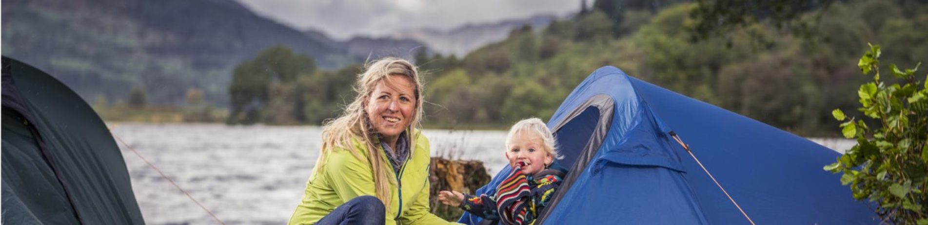 blonde-woman-holding-baby-at-blue-tent-entrance-at-edge-of-loch-chon