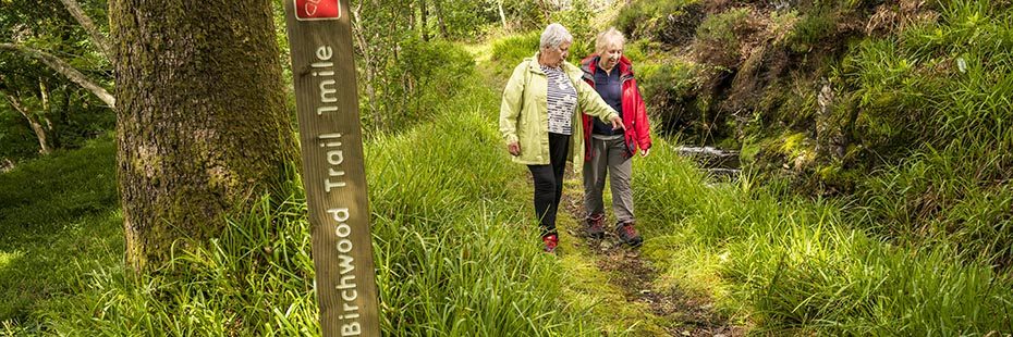 two-elderly-ladies-dressed-in-colourful-outdoor-gear-walking-on-path-in-lush-green-forest-at-ardentinny