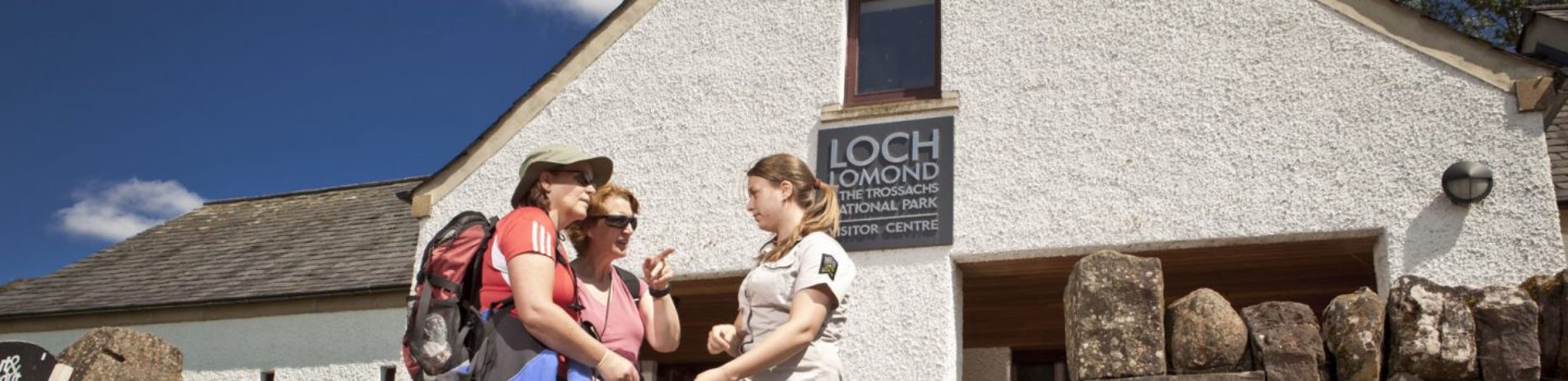 national-park-ranger-advising-two-women-walkers-in-front-of-balmaha-visitor-centre-on-blue-skies-summer-day