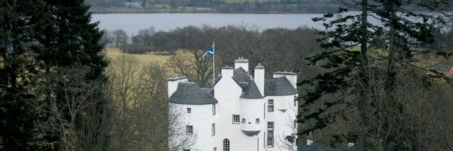 white-castle-edinample-with-scottish-blue-and-white-flag-above-the-slate-roof-surrounded-by-bare-forests-and-loch-earn-visible-behind