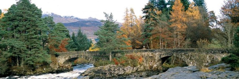 killin-village-stunning-falls-of-dochart-next-to-ancient-stone-bridge-trees-surrounding-it-and-high-mountains-in-distance-with-a-smatter-of-snow-on-top