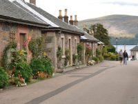 luss-pier-road-with-loch-lomond-and-tourists-in-the-distance-and-stone-houses-with-flowers-lining-the-street