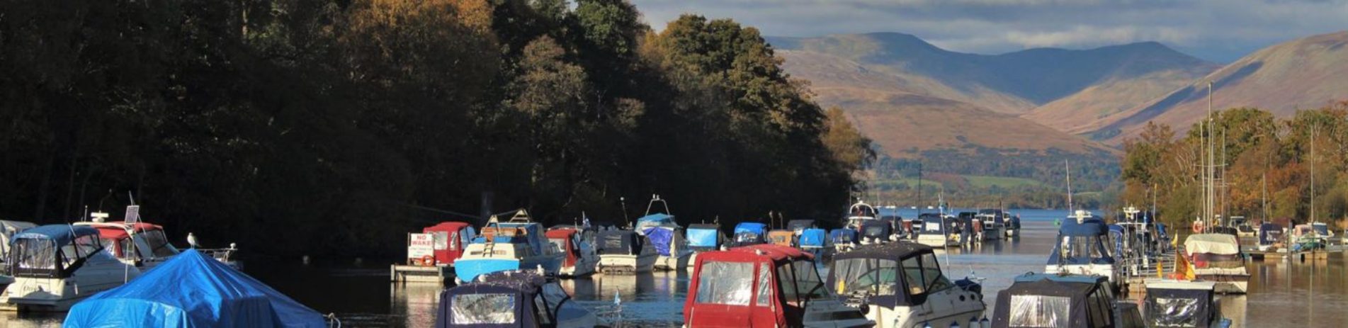 colourful-boats-on-river-leven-in-balloch-with-loch-lomond-opening-behind-and-luss-hills-half-covered-in-shadows