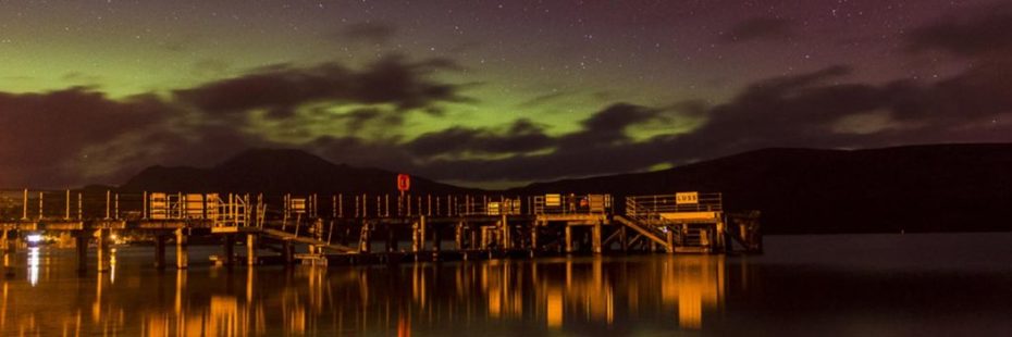 luss-pier-lit-up-in-the-night-northern-lights-green-and-purple-visible-behind-above-ben-lomond-and-hills