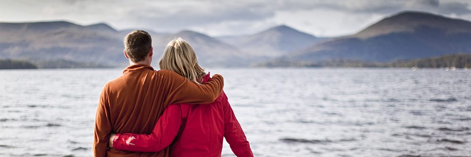 couple-holding-each-other-affectionately-looking-into-distance-admiring-stunning-landscape-of-loch-lomond-and-luss-hills-at-milarrochy-bay