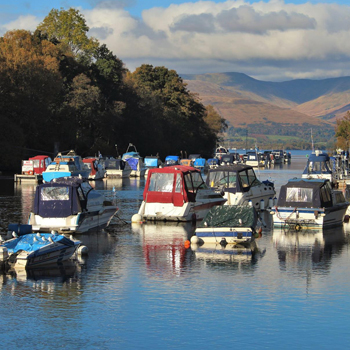 colourful-boats-on-river-leven-in-balloch-with-loch-lomond-opening-behind-and-luss-hills-half-covered-in-shadows