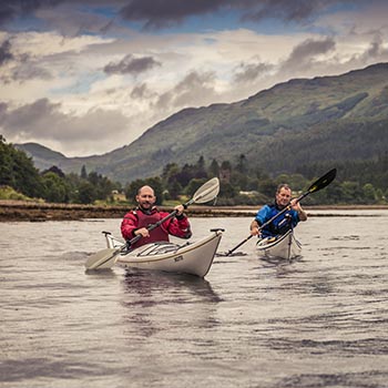 two-men-kayaking-one-in-red-and-the-other-in-blue-at-ardentinny-with-forested-hills-behind