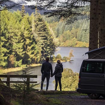 young-couple-holding-hands-admiring-stunning-loch-and-forests-view-on-high-ground-next-to-campervan-at-three-lochs-forest-drive-site