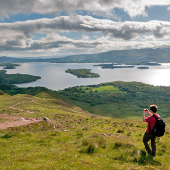 young-man-in-red-t-short-and-backpack-talking-photo-of-stunning-loch-lomond-and-islands-from-the-slopes-of-conic-hill
