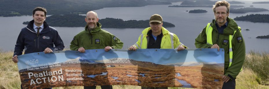 peatland-action-group-of-four-including-national-park-staff-smiling-for-camera-up-on-hill-with-loch-lomond-and-islands-visible-behind