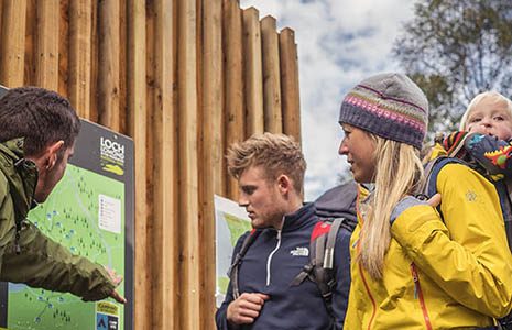 national-park-ranger-in-green-jacket-pointing-on-map-to-young-couple-with-baby-at-loch-chon-campsite