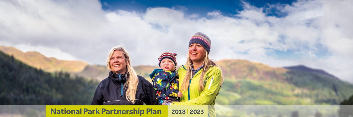 two-blonde-women-holding-baby-and-looking-into-distance-at-loch-chon-with-text-banner-underneath-reading-national-park-partnership-plan-two-thousand-eighteen-two-thousand-twenty-three