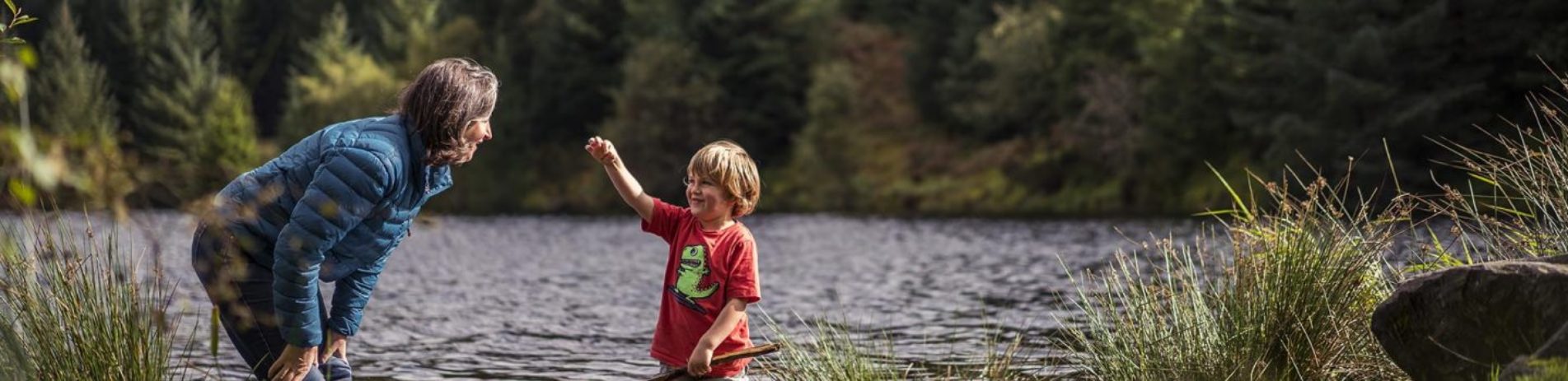 woman-in-blue-jacket-stopping-and-looking-at-young-boys-hand-they-are-both-in-water-ankle-deep-three-lochs-forest-drive