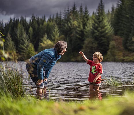woman-in-blue-jacket-stopping-and-looking-at-young-boys-hand-they-are-both-in-water-ankle-deep-three-lochs-forest-drive