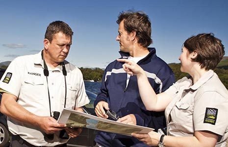 two-rangers-talking-to-member-of-the-public-and-advising-him-using-map-at-milarrochy-bay-on-summer-day-boat-launching-behind