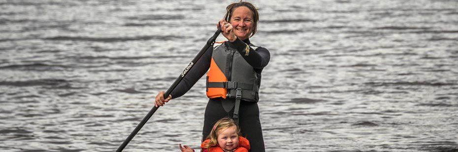 woman-in-life-vest-and-wetsuit-with-very-young-daughter-paddleboarding-happily-on-loch